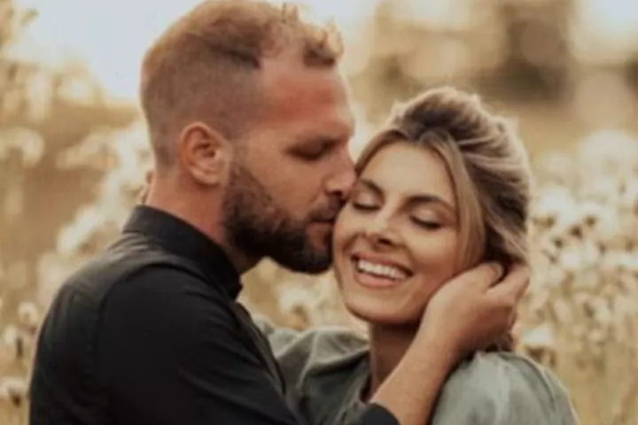 Couple enlacé dans un champ, l'homme embrasse la joue de sa compagne qui sourit